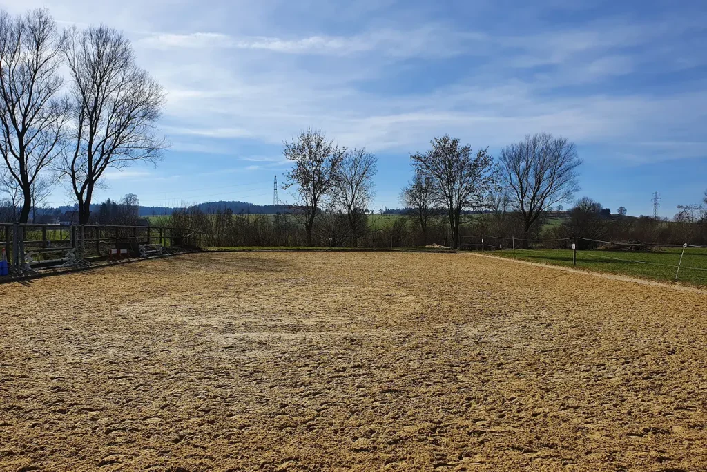 Eingezäunter Outdoor-Reitplatz mit hellem Sandboden vor einer Hügellandschaft unter blauem Himmel.